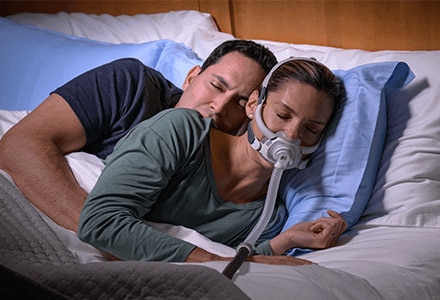 Uma mulher a dormir tranquilamente de lado com a máscara PAP AirFit F40 da ResMed e o seu parceiro enrolado atrás de si.